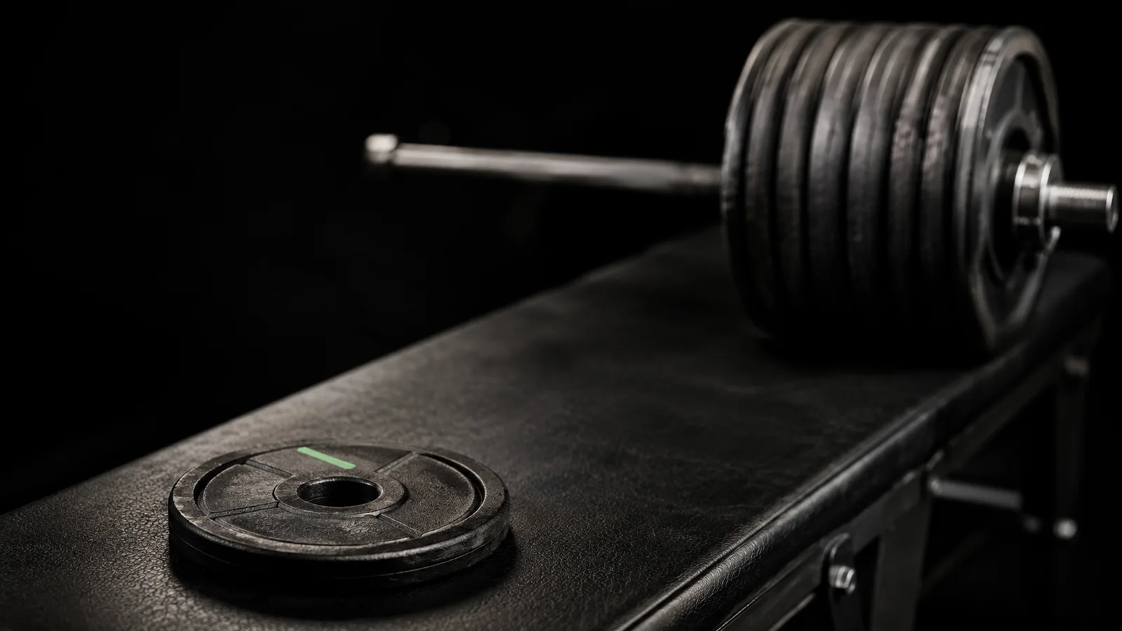A single black weight plate with a green accent stripe resting alone on a dark leather bench, with a heavily loaded barbell extending into soft focus in the background, visually contrasting a four-week starting point with a twelve-week result