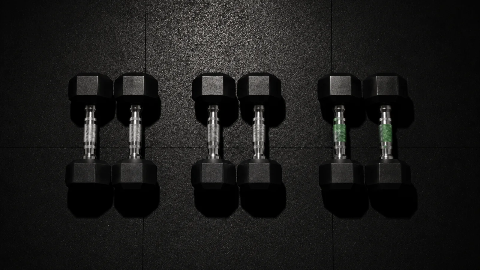 Three matching pairs of hex dumbbells arranged in a row on a dark textured gym floor under an overhead light, one pair with green-accented grips, symbolizing a small-group semi-private training session
